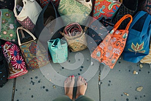 feet surrounded by assorted fabric totes on a sidewalk