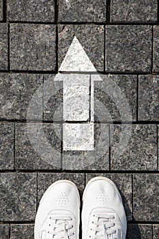 Feet white sneakers standing on a cobblestone pavement in front the white direction sign to go ahead