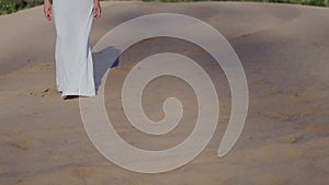 Feet of woman walking on beach dunes in white dress while sun drops shadow slow motion front view