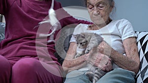Feline therapy. Mature daughter visits senior mother in nursing home, plays with her and kitten on couch. Old woman