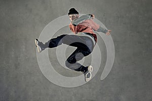 Fellow with tattooed face, beard. Dressed in hat, colorful jumper, black pants and sneakers. Jumping, dancing on gray
