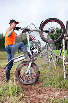 Fellow with trial mototrcycle training in countryside area, raising bike on rear wheel, leaning against wooden fence