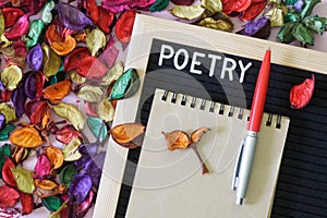 Felt board with the inscription poetry, a notebook and a fountain pen next to a lot of multi-colored dry leaves and petals. The