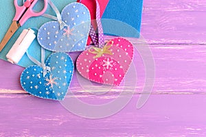 Felt hearts crafts decorated with beads and snowflakes, scissors, thread, needles, felt sheets on lilac wooden background