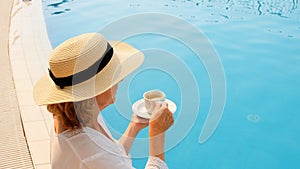 female 50 years old having breakfast by the pool in a straw hat wearing a white dress. woman sitting by the pool with a
