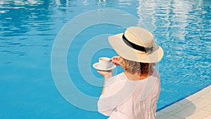 female 50 years old having breakfast by the pool in a straw hat wearing a white dress. woman sitting by the pool with a