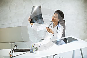 Female African American doctor wearing white coat with stethoscope sitting behind desk in office and looking x-ray image