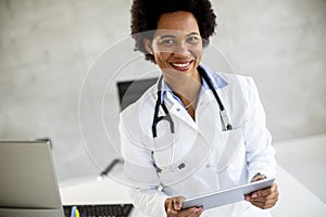 Female African American doctor wearing white coat with stethoscope standing by desk in office and holding digital tablet