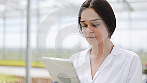 Female agricultural enginee with tablet computer walking along greenhouse, examining state of plants and passing by farm