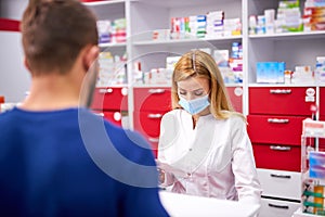 Female apothecary in protective medical mask and young man customer buying drug at drugstore