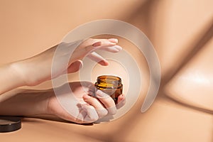 Female applying a collagen firming cream to a hand. Open cream jar on beige background. Cosmetics mockup. Trendy colors