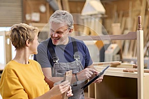 Female Apprentice Learning From Mature Male Carpenter With Digital Tablet  In Furniture Workshop