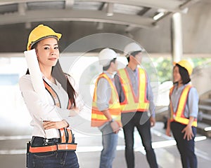 Female architect or engineer holding blue print paper, standing in front of her team smiling and looking at camera