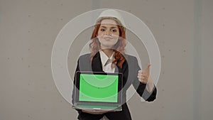 A female architect holds a laptop screen facing forward, giving a thumbs up. Portrait of a business woman in a hardhat