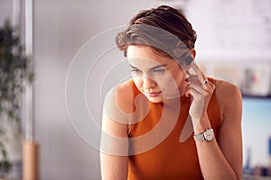 Female Architect In Office Standing At Desk Taking Phone Call On Wireless Earpiece