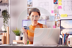 Female Architect In Office Working At Desk Making Online Purchase Using Credit Card On Laptop