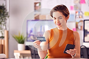 Female Architect In Office Working At Desk Making Online Purchase Using Credit Card On Mobile Phone