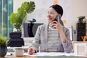 Female Architect Working In Office Sitting At Desk Talking On Mobile Phone