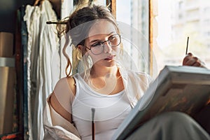 Female artist painting on canvas in her art studio sitting next to the window. A woman painter with transparent eyeglasses