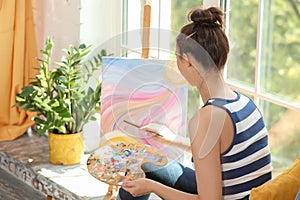 Female artist painting picture while sitting on windowsill in workshop