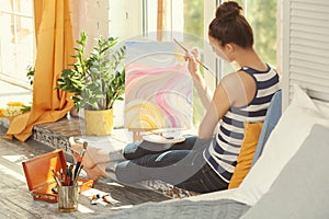 Female artist painting picture while sitting on windowsill in workshop