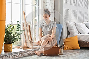 Female artist painting picture while sitting on windowsill in workshop