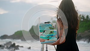 Female artist paints colorful seascape on canvas at stormy beach with waves crashing. Tattoo visible on arm as adds