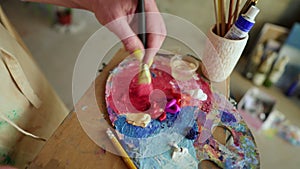 Female artist with thick paintbrush is mixing different coloured oil paints on palette - purple, red, yellow. Close up