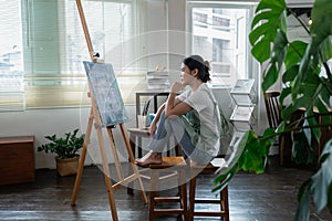 Female artist thinking and hold paintbrush while looking at canvas and pondering about paintin