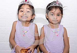 Female asian identical twins sitting on chair with white background. Wearing purple dress and accessories like necklace, crown