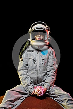 female astronaut in spacesuit and helmet sitting on planet with flower