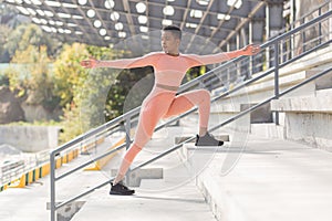 Female athlete doing fitness in a pink sports suit near the sports stadium, African American performs fitness exercises and
