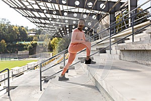 Female athlete doing fitness in a pink sports suit near the sports stadium, African American performs fitness exercises and