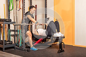 Female athlete exercising on bar bench press in a gym with help of coach