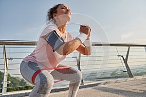 Female athlete exercising with elastic band on the city bridge. Sports woman doing body weight training outdoor early in the