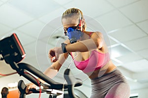 Female athlete with oxygen mask checking time while exercising with exercise bike in fitness studio