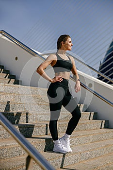 Female athlete in sportswear have a rest after jogging outside standing on steps and looks away