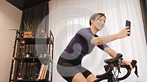 Female athlete takes selfies on smartphone while cycling on exercise bicycle. Woman is training on smart bike trainer. Indoor cycl