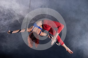 Female athletic, sexy and flexible aerial circus artist with redhead on aerial straps on black background