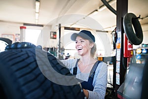 Female auto mechanic changing tieres in auto service. Beautiful woman holding tire in a garage, wearing blue coveralls.