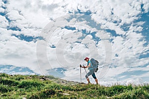 Female backpacker climbing to the mountain top using trekking poles with bright cloudscape background. Active vacation spending by