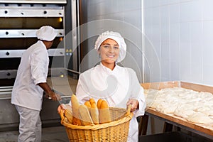 Female baker holding basket of hot baguettes in the kitchen of bakery