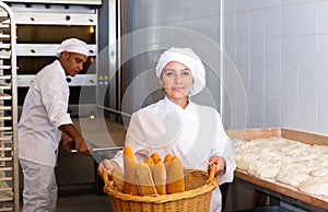 Female baker holding basket of hot baguettes in the kitchen of bakery