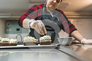 Female baker wearing glove and black apron decorating chocolate frosted cupcakes with sprinkles. Professional baking