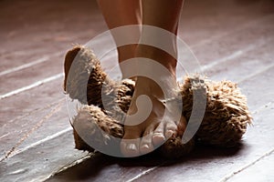 female bare feet stepping on a brown teddy bear on the old floor in a dark room, a toy on the floor with legs laid