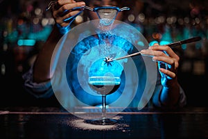 Female bartender adding to a brown cocktail and pour on a flamed badian on tweezers a powdered sugar in the blue light photo
