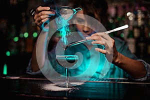 Female bartender pouring on the brown cocktail and on a flamed badian on tweezers a powdered sugar in the green light photo