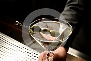 Female bartender serving transparent cocktail based on martini decorated with olives on the skewer