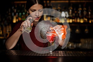 Female bartender sprinkling a cocktail glass with Aperol syringe cocktail with a peated whisky and making a smoky note on the bar