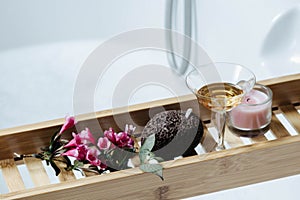 Female bathroom - romantic bath with natural pumice, glass of white wine, pink flowers and candle on shelf for a bathtub